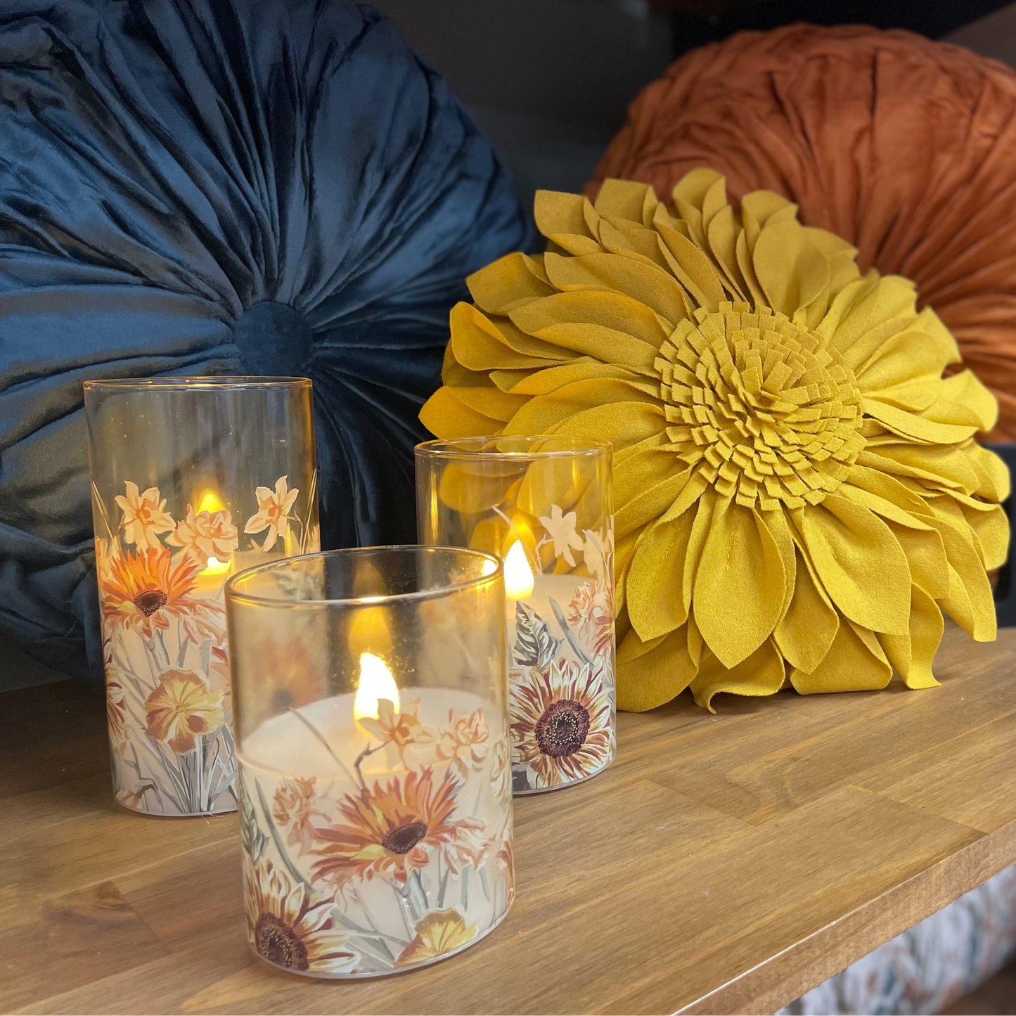 Decorative candles with floral designs on a wooden surface with decorative pillows and plants in the background.