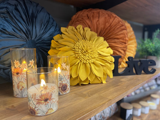 Decorative candles with floral designs on a wooden surface with decorative pillows and plants in the background.